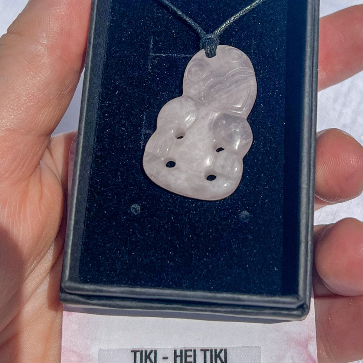 Hand holding a tiki pendant in a display case with 'Tiki - Hei Tiki' text below.