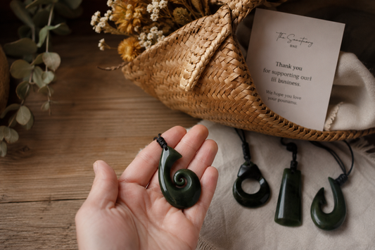 Hand holding an authentic New Zealand Greenstone pendant with other pounamu necklaces displayed on a rustic wooden table.