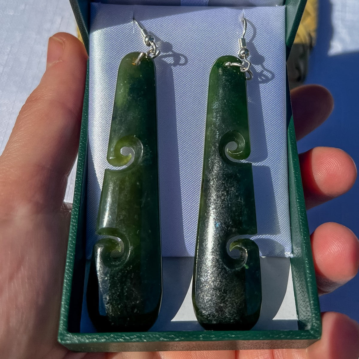 Hand-carved NZ greenstone double koru drop earrings displayed in a green gift box, held in hand to show scale — authentic pounamu, 70mm