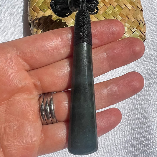 Hand holding NZ Greenstone pendant with a woven kete bag in the background