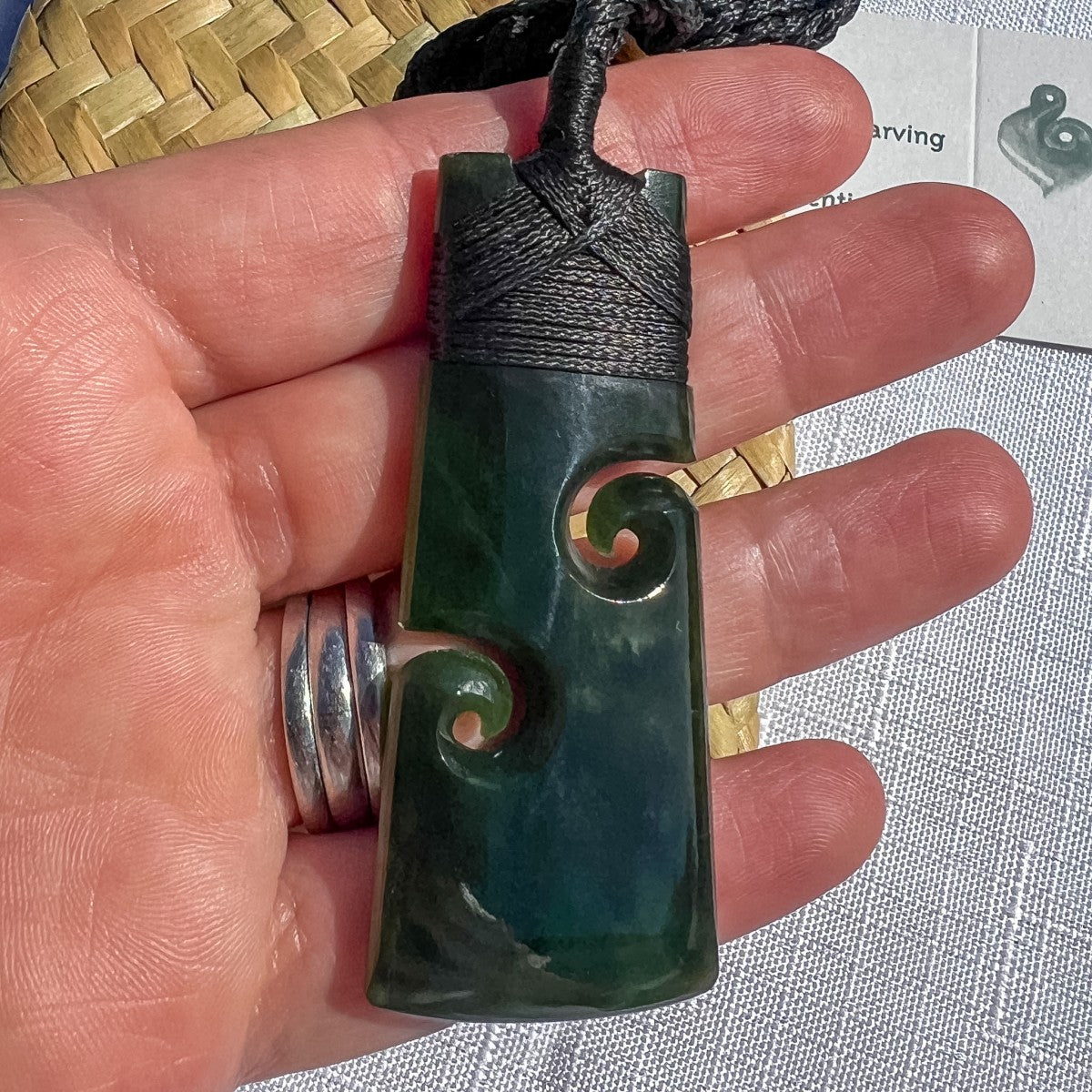 Hand-carved NZ greenstone toki koru pendant 60mm on black cord, held in hand to show scale — deep green pounamu with double koru carving detail