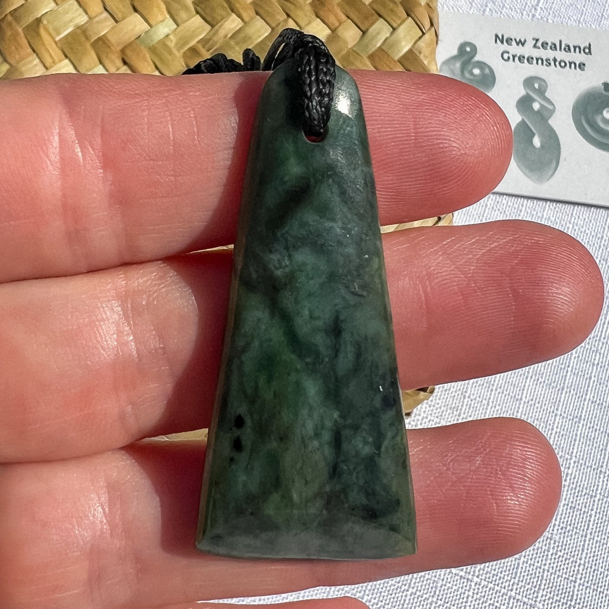 Hand-carved NZ greenstone toki pendant 50mm on black cord, held in hand to show scale — deep green pounamu with natural mottling, drilled at top