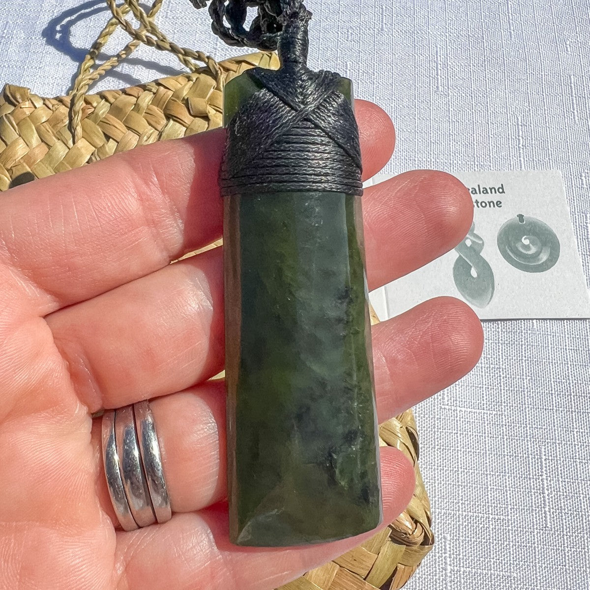 Hand-carved NZ greenstone toki pendant 70mm on black cord with woven binding, held in hand to show scale — authentic pounamu