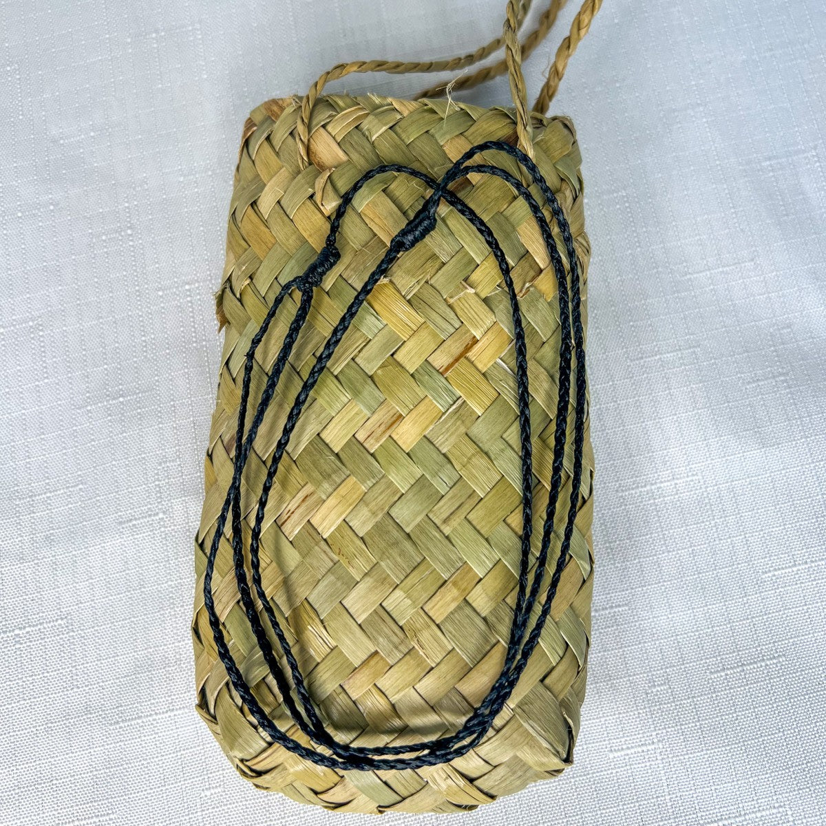 Braided black slip knot cord for greenstone pendant, displayed neatly coiled on top of a traditional woven flax kete bag.