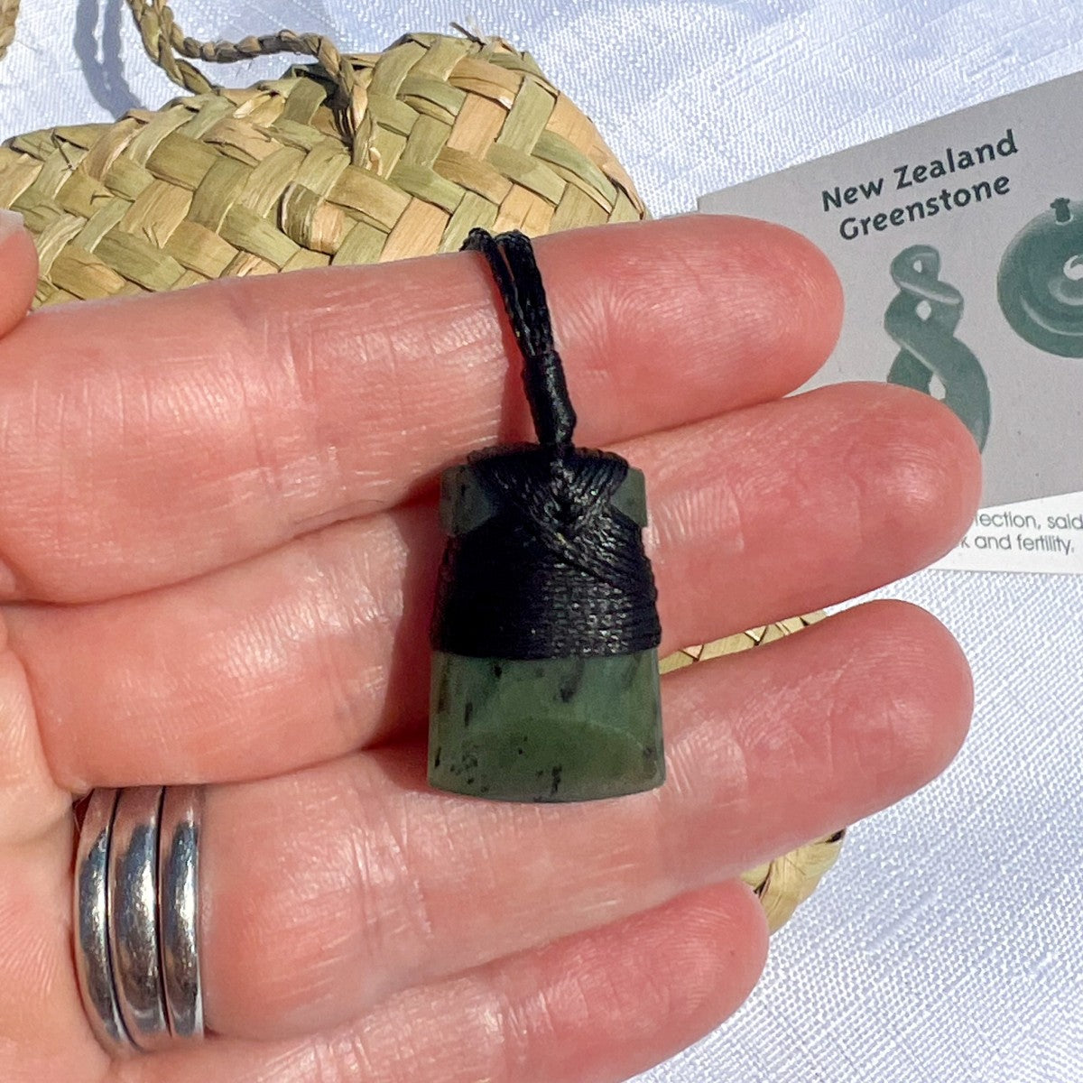 Greenstone pendant on a black cord held between fingers with a woven mat and card in the background.