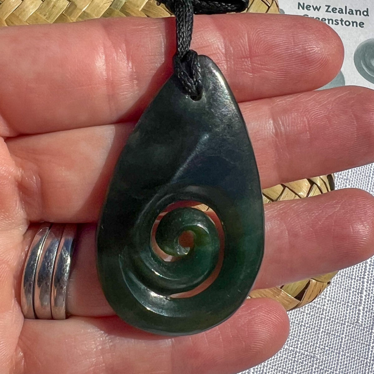 A dark green teardrop-shaped carved pendant with a spiral motif, held in a person's hand. The pendant is made of polished stone and hangs from a black cord, with a silver ring on the person's finger.