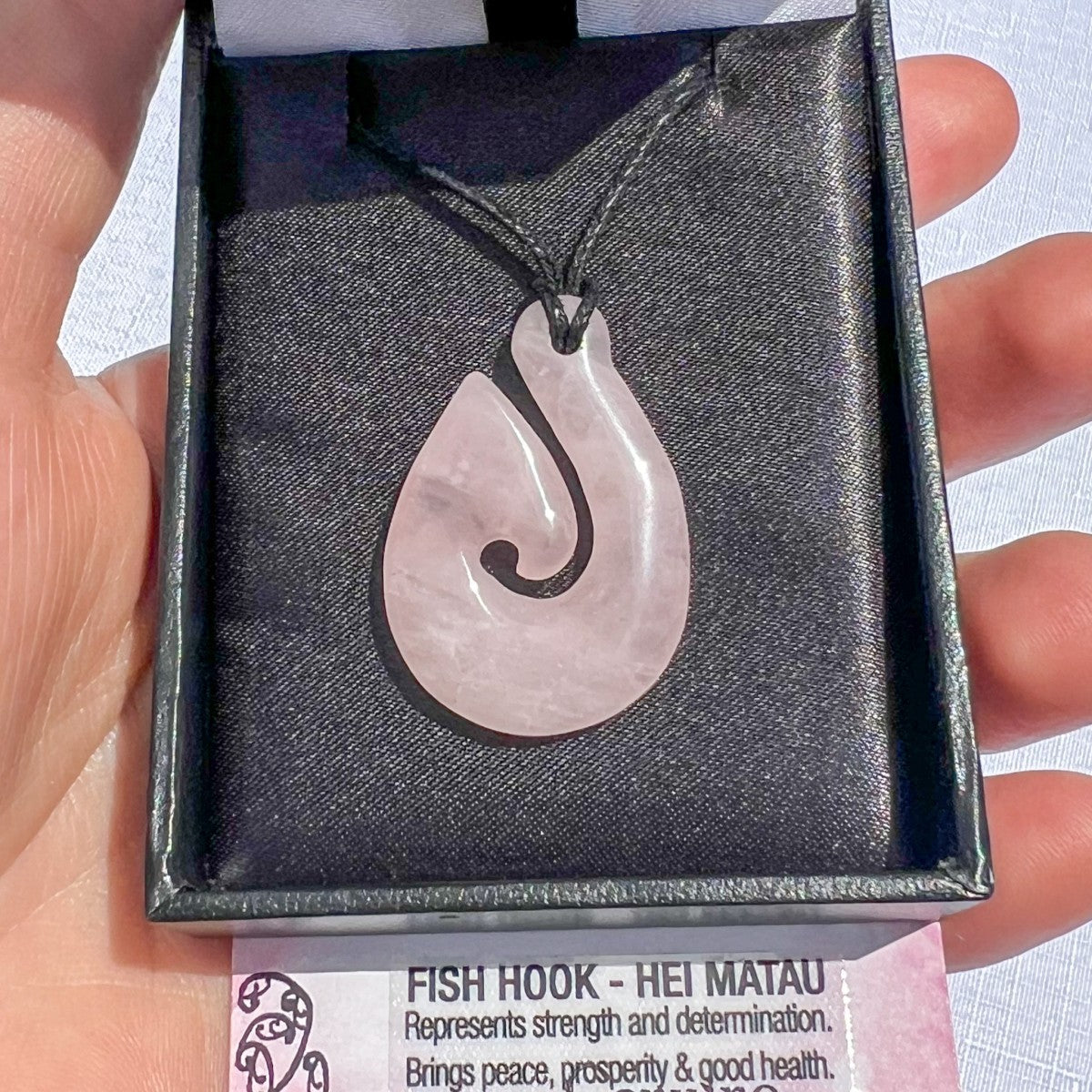 Rose quartz Hei Matau (fish hook) pendant necklace on a black cord, symbolising love, strength, and safe travel — hand-carved in New Zealand.